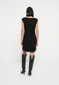 Femme aux cheveux courts noirs portant une robe noire texturée sans manches au genou et des bottes noires à talons hauts, se tenant face à un mur blanc.
