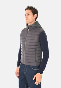 Uomo che indossa un gilet grigio imbottito senza maniche con zip, sovrapposto a una camicia a maniche lunghe blu navy e jeans blu, rivolto a sinistra in luce neutra.