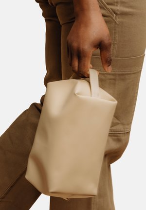 Hånd holder en beige rektangulær veske i det korte håndtaket, med brune bukser synlige i bakgrunnen.