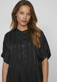 Vestito nero con strisce verticali, maniche corte, chiusura frontale con bottoni, tessuto leggero, vestibilità comoda. Nota lucentezza della trama e scollatura a barca.
