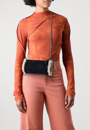 Vrouw draagt oranje top met patroon en lange mouwen en koraalrode broek met zwarte crossbodytas met nepbontbies en kettingriem.