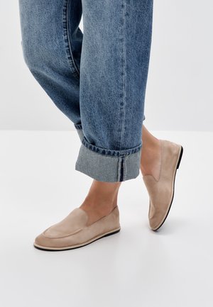Pieds portant des mocassins beige à enfiler, associés à un jean en denim bleu retroussé, sur un fond blanc uni.