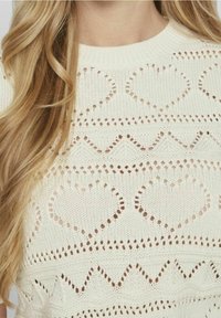 pletený svetr v krémové barvě s otevřeným vzorem, který zobrazuje srdce a zigzagové designy, kulatým výstřihem a texturovanou přízí.