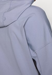 Lichtblauwe capuchontrui gemaakt van zacht, rekbaar materiaal met zichtbare naden en een licht gestructureerde afwerking, voorzien van een enkele zijzak.