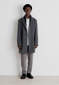 Cappotto di lana grigio con due bottoni e colletto a punta, indossato sopra una camicia bianca, abbinato a pantaloni grigi chiari e scarpe nere.