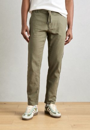 Jasnooliwkowe spodnie wykonane z teksturowanej tkaniny z przesuwną talią. Połączone z jasnymi butami sportowymi z zielonymi akcentami.