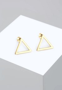 Zlaté trojuholníkové náušnice s otvoreným dizajnom a hladkým povrchom, vystavené na bielom geometrickom podstavci. Minimalistický štýl.