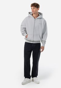 Lys grå zip-up hættejakke med en frontlomme, der har et diskret logo. Sammen med mørke jeans og hvide sko, står personen foran en simpel baggrund.
