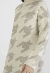 Pull en tricot à motif pied-de-poule crème et gris clair porté par une personne, montrant le torse supérieur et le bras droit avec la main détendue.