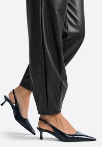 Melnas lakotas ādas sīkrēka papēdītes ar smailu purngalu un mazu kitten papēdi. Gluda tekstūra ar regulējamu sprādzi aizmugurējā siksniņā.