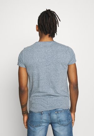 Mężczyzna o dredach, ubrany w szarą koszulkę z krótkim rękawem i niebieskie dżinsy, stoi twarzą do gładkiej białej ściany, widziany od tyłu.