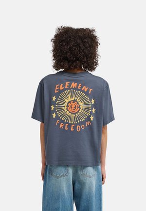 Persoon met krullend haar die een donkerblauw T-shirt draagt met een geel- en oranje zon- en sterrenmotief en de tekst "Element Freedom" op de rug.