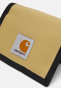 Yellow canvas wallet with black trim, featuring a white label with a circular orange logo and the text "carhartt." Textured surface.