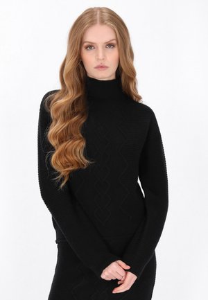 Jonge vrouw met lang golvend blond haar, gekleed in een zwarte, getextureerde trui met hoge hals, staand met gevouwen handen tegen een effen witte achtergrond.