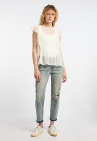 Weißes Rüschenshirt mit einem durchsichtigen, gepunkteten Overlay, kombiniert mit hellblauen, verwaschenen Jeans und weißen Sneakers.