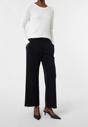 Femme portant un haut texturé blanc à manches longues, un pantalon large noir et des escarpins noirs à bout pointu et à bride arrière, debout les mains dans les poches.