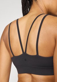Μαύρο αθλητικό σουτιέν με διπλές τιράντες, λεία υφή, εφαρμοστό σχέδιο και διακριτική λεπτομέρεια με λογότυπο στο πίσω μέρος. Η πίσω όψη αναδεικνύει το σχέδιο με τις τιράντες.