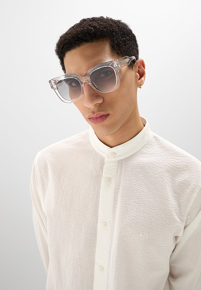 Gafas de sol grandes y transparentes con lentes gris degradado; montura transparente. Camisa blanca texturizada con cuello alzado y frente de botones.