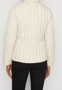 Pikowana kremowa kurtka z dopasowaną talią, długimi rękawami i wysokim kołnierzem. Posiada pionowe przeszycia i gładką teksturę.