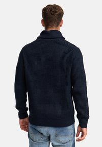 Granatowy sweter z dzianiny z wysokim kołnierzem, z teksturowanym wzorem waflowym i ściągaczami. Stylizowany z casualowymi niebieskimi dżinsami.