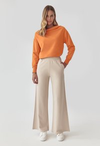 Hanorac portocaliu cu o croială lejeră și gât rotund, asortat cu pantaloni bej cu croială largă. Adidași albi completează ținuta.