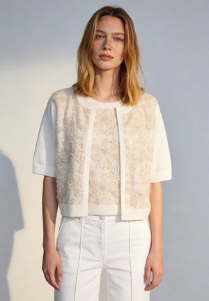 Junge Frau mit schulterlangen blonden Haaren trägt eine weiße, kurzärmelige bestickte Strickjacke und weiße, hoch taillierte Hosen.