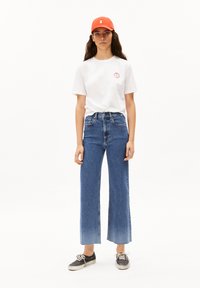 Camiseta de algodón blanca con un pequeño logo redondo, vaqueros azules de cintura alta con piernas anchas y una gorra naranja. Zapatillas negras con suelas blancas.