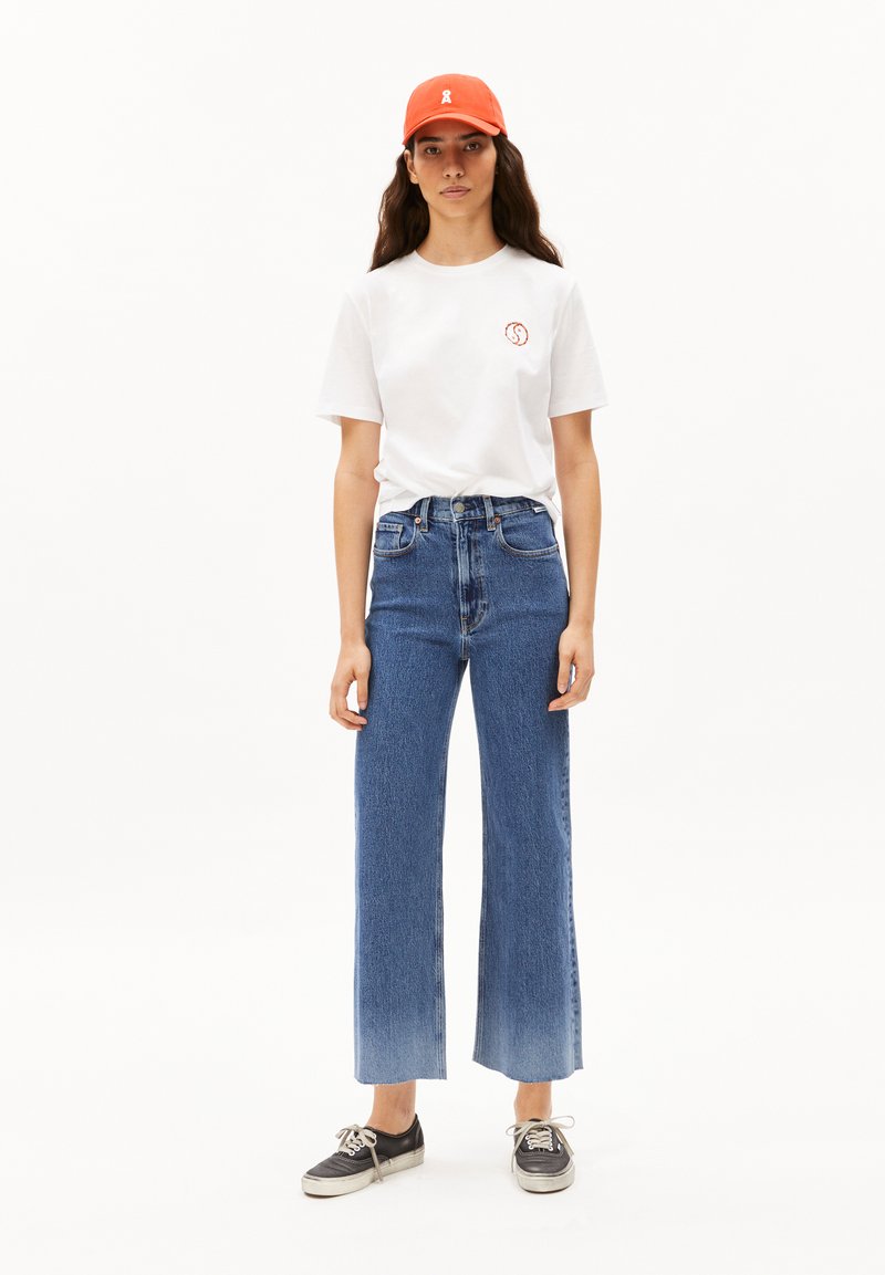 Camiseta de algodón blanca con un pequeño logo redondo, vaqueros azules de cintura alta con piernas anchas y una gorra naranja. Zapatillas negras con suelas blancas.