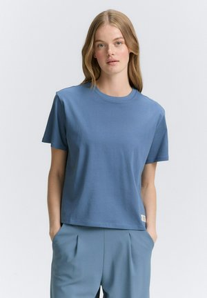 T-shirt à manches courtes et col rond en coton doux, bleu clair. Présente une coupe décontractée et une petite étiquette de marque à l'ourlet. Texture lisse.