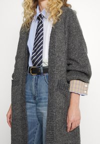 Grå, ribbad cardigan med långa ärmar, lager-på-lager över en ljusblå randig skjorta och en mörk randig slips, kombinerad med höga blå jeans.