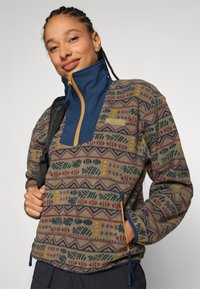 Fleece pullover med et mønstrets olivengrønt krop og marineblå krave, en halvlukket front og gyldne detaljer. Inkluderer sidelommer med lynlås.
