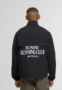 Uomo in giacca nera con la scritta bianca "Sunday Running Club" sulla schiena, in piedi su uno sfondo chiaro e uniforme.