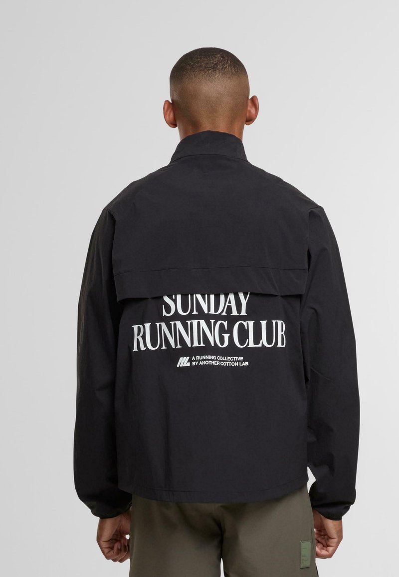 Uomo in giacca nera con la scritta bianca "Sunday Running Club" sulla schiena, in piedi su uno sfondo chiaro e uniforme.
