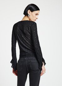 Top noir à manches longues en tissu texturé, avec des poignets évasés. Porté avec un jean foncé, mettant en valeur une silhouette élégante et ajustée.