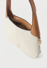 Beżowa i brązowa torba na ramię z wzorzystej tkaniny, ze skórzanymi wykończeniami, złotym zamkiem błyskawicznym i okrągłą złotą zawieszką z logo na białym tle.