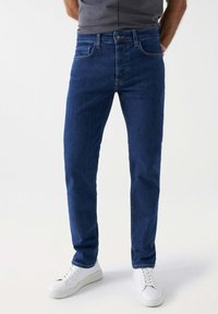 Jeans de mezclilla azul oscuro con un corte ajustado, cinco bolsillos y un cierre de botón clásico. Combinados con zapatillas blancas sobre un fondo liso.