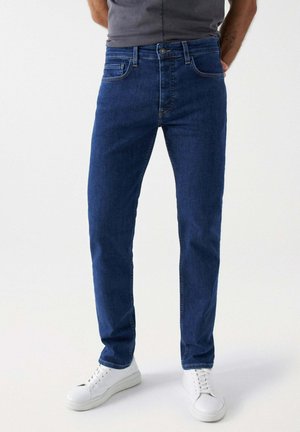 Jeans de mezclilla azul oscuro con un corte ajustado, cinco bolsillos y un cierre de botón clásico. Combinados con zapatillas blancas sobre un fondo liso.