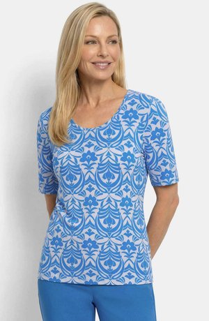 Femme souriante, portant une chemise à manches courtes à motif floral bleu et blanc et un pantalon bleu uni sur un fond uni.