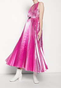 Femme portant une robe midi rose brillante et plissée sans manches avec une ceinture à la taille, associée à des bottines blanches, debout devant un fond blanc.