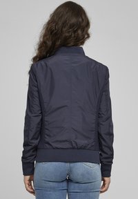 Námořnická modrá bomber bunda vyrobená z hladkého nylonu, s žebrovanými manžetami a lemem a texturovanými bočními panely. Pohled zezadu ukazuje minimalistický design.