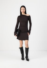Hnědress s odhalenými rameny a dlouhými rukávy, se zvýrazněnou siluetou, v kombinaci s černými vysokými botami s špičatou špičkou a malou černou kabelkou.