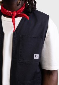 Svart ermeløs vest med glidelås foran, brystlomme og logo-patch; brukt over en hvit T-skjorte og paret med et rødt bandana.