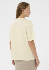 Femme aux cheveux longs portant une chemise beige à manches courtes et une jupe beige, de profil contre un fond blanc uni.