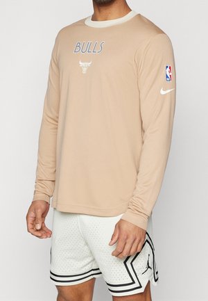 Homme portant un t-shirt à manches longues beige des Chicago Bulls avec les logos NBA et Nike, et un short de basket blanc avec une bordure noire et le logo Jordan.