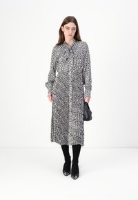 Schwarz-weiß gemustertes Kleid mit langen Ärmeln, Kragen und Taillengürtel. Das Modell trägt schwarze Strumpfhosen und hohe Schuhe und hält eine kleine schwarze Tasche.