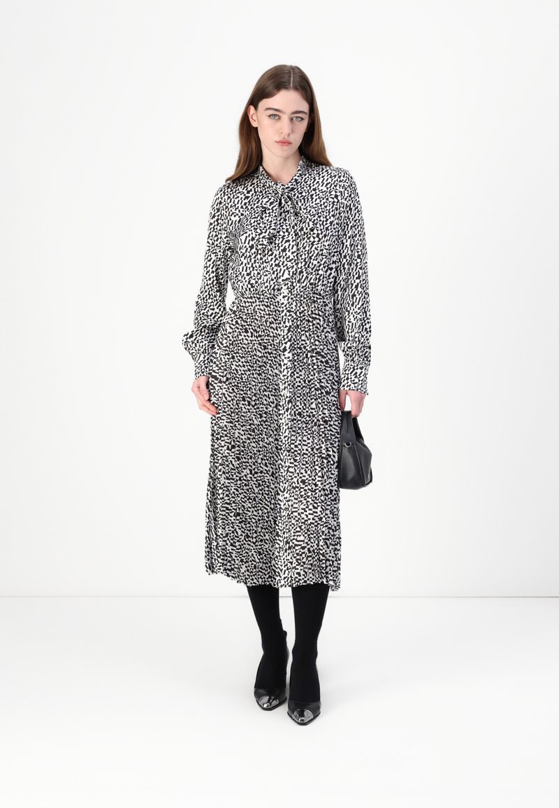 Schwarz-weiß gemustertes Kleid mit langen Ärmeln, Kragen und Taillengürtel. Das Modell trägt schwarze Strumpfhosen und hohe Schuhe und hält eine kleine schwarze Tasche.