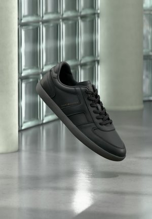Schwarzer Tommy Hilfiger Sneaker mit Schnürsenkeln, der über einem glänzenden Betonboden in der Nähe einer Glassteinwand und einer weißen Säule schwebt.