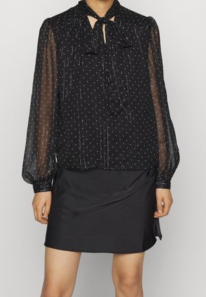 Woman wearing a black sheer long-sleeve blouse with white polka dots and a bow tie neckline paired with a simple black skirt.