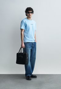 Homme portant un t-shirt bleu clair et un jean, tenant une mallette noire, portant des lunettes de soleil noires, debout contre un mur gris clair uni.