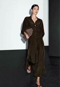 Ensemble deux pièces en vert olive foncé, en tissu soyeux, comprenant une blouse boutonnée et un pantalon large fluide. Sac à main marron et sandales à lanières.